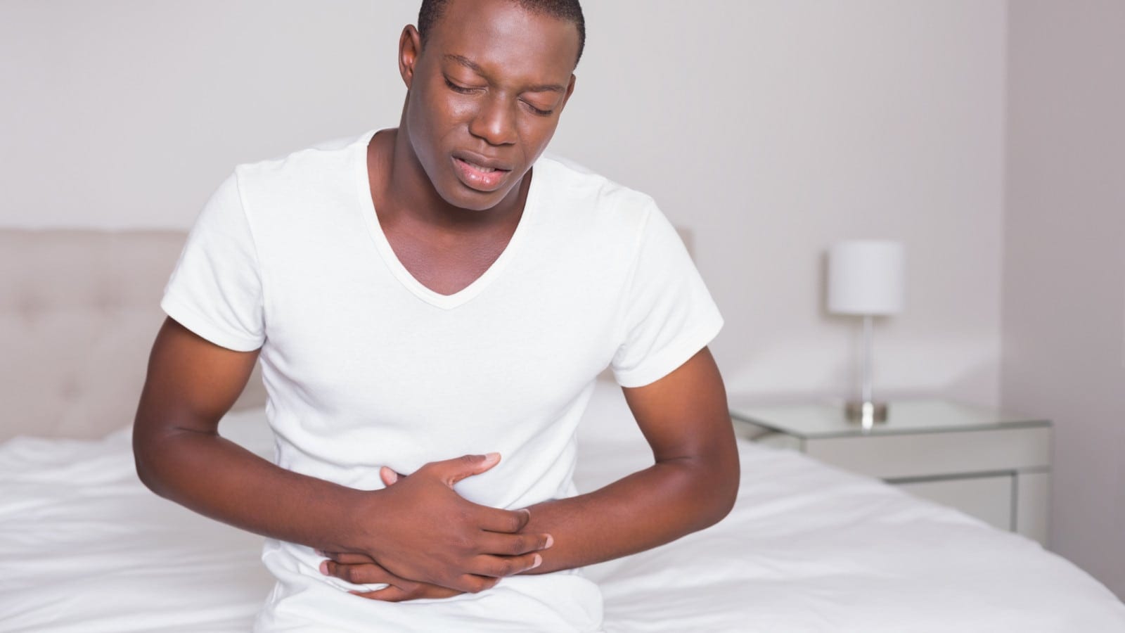 Wincing man sitting on the edge of his bed with a stomach cramp at home in bedroom